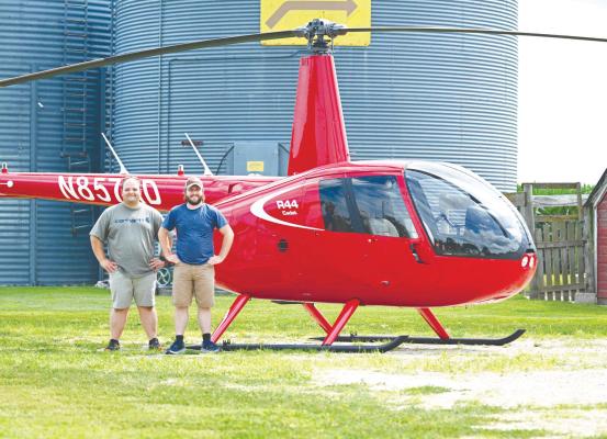 ST. PAUL native Nathan Bauer (right) is pictured with his fellow flight instructor Izak Snell in front of an R44 Cadet helic opter, which the two landed on Bauer’s family farm near Cushing on Saturday. Photo by Michael Happ