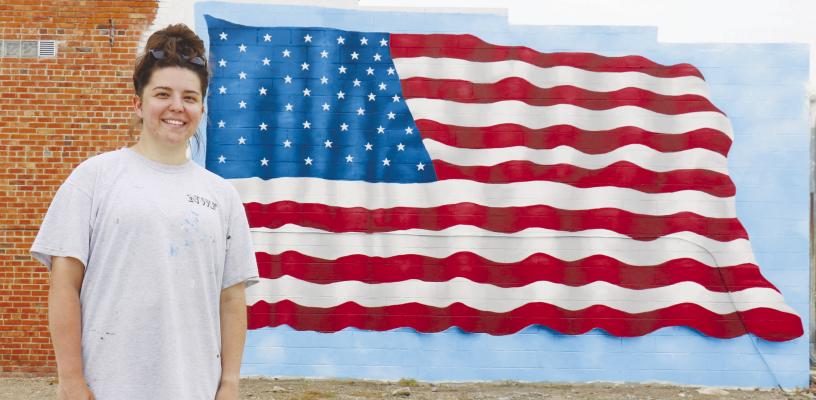 NEELY TUBBS, a 2016 graduate of St. Paul High School, is pictured in front of an American flag mural that she recently painted on the west wall of Luke’s Shoe and Saddle Shop in downtown Wolbach. The Wolbach mural was the fourth created by the artist. Photos by Michael Rother